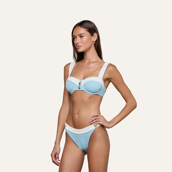 A woman poses with one hand on her hip against a plain light background, wearing the Jean Bikini Bottom—a light blue and white textured bouclé swim bikini featuring buttons on the top and a high-leg cut bottom.