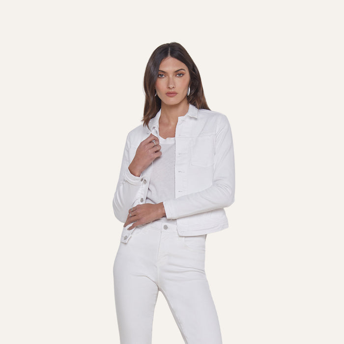 A woman stands against a plain background, wearing the Celine Jean Jacket in optic white with a relaxed fit, layered over a matching shirt and white jeans. She has straight brown hair and a neutral expression, with one hand touching the jacket near the collar.