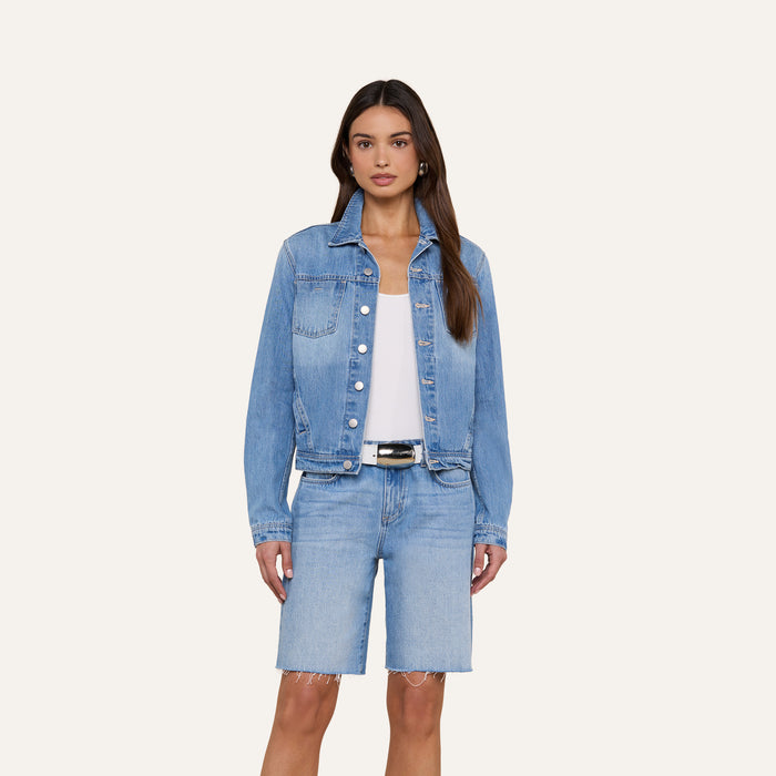 A woman stands against a plain light background wearing the Celine Jean Jacket from L’AGENCE in a light denim wash, styled over a white top and matching knee-length denim shorts.