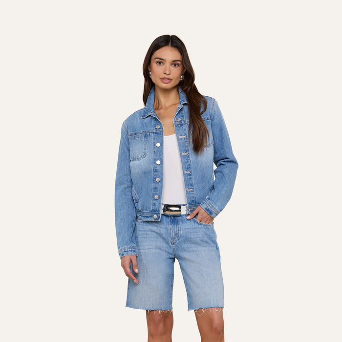 A woman wearing the Celine Jean Jacket in a light denim wash with matching knee-length denim shorts, a white top, and a black belt stands with one hand in her pocket against a plain background.