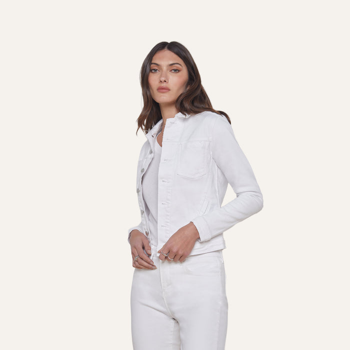 A woman with long brown hair wears the Celine Jean Jacket in optic white, featuring a relaxed fit, layered over a light top and matching white jeans, standing against a plain off-white background.