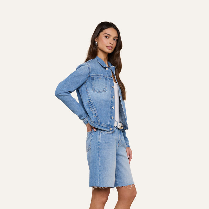 A woman stands in a light denim wash Celine Jean Jacket and matching slim fit, knee-length denim shorts against a plain, light background. She has long brown hair and is looking slightly to the side.