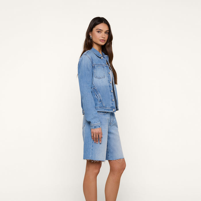 A woman stands sideways, wearing a slim fit light denim wash Celine Jean Jacket and matching knee-length denim shorts against a plain white background.