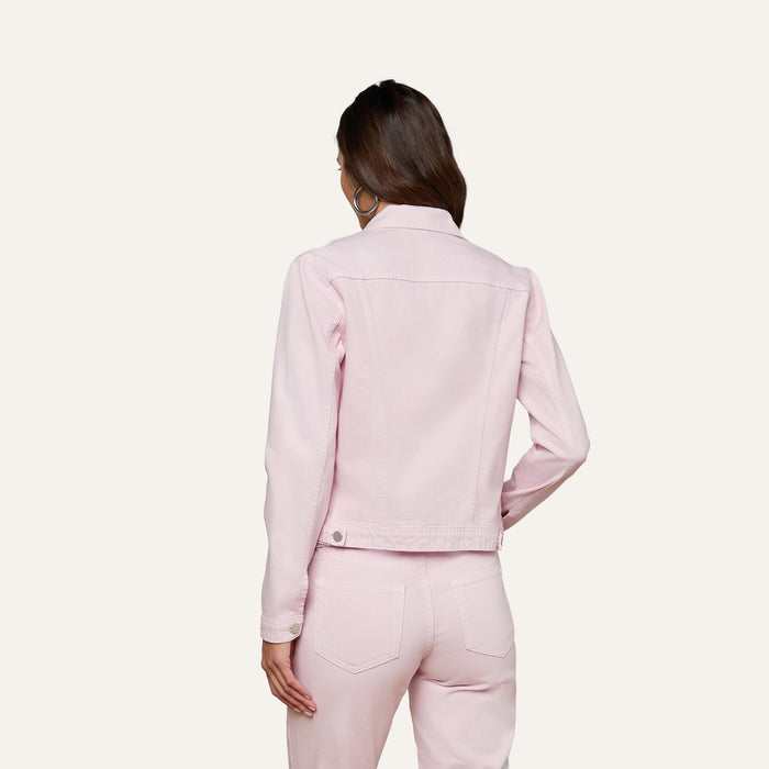 A woman stands facing away, wearing a pastel lilac Celine Jean Jacket by L’AGENCE with matching pants. Her right hand rests on her hip, and her brown hair falls over her shoulders against a plain light background.