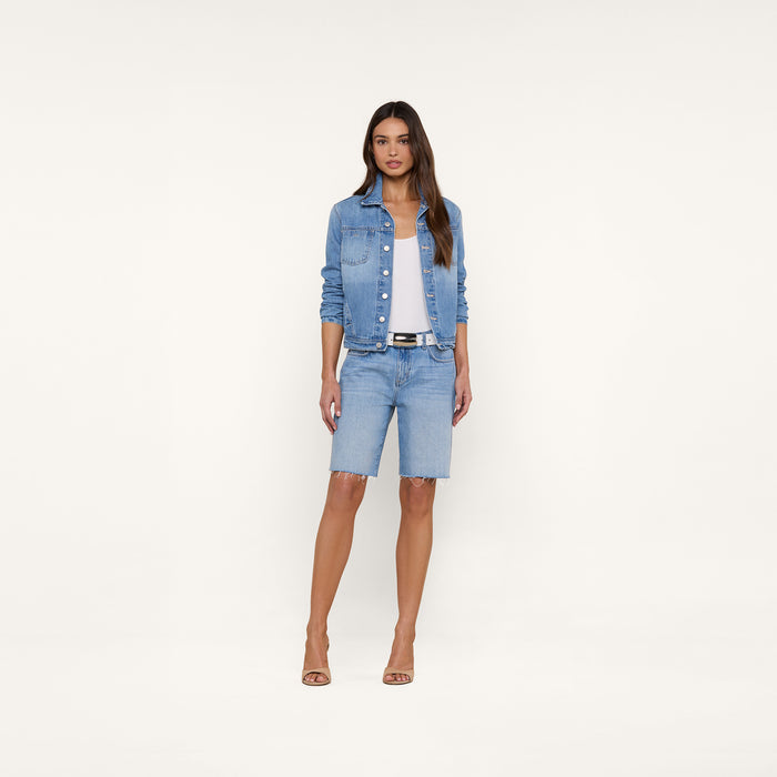 A woman stands facing forward, wearing the Celine Jean Jacket by L’AGENCE in a light denim wash, a white top, slim fit light blue knee-length denim shorts, a black belt, and beige heels against a plain white background.