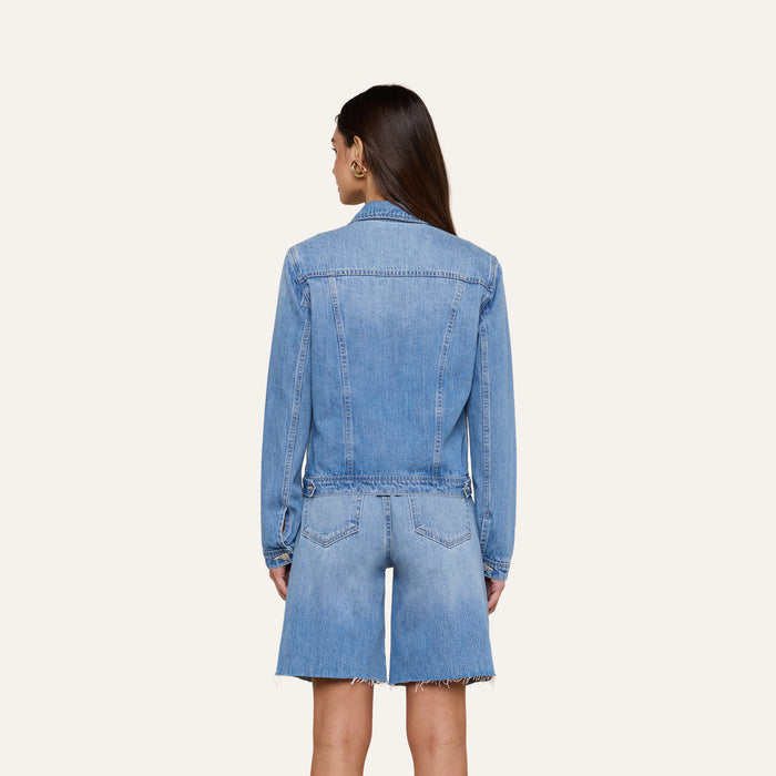 A woman with long brown hair stands facing away, wearing a slim fit Celine Jean Jacket by L’AGENCE in a light denim wash and matching knee-length denim shorts with frayed hems against a plain light background.