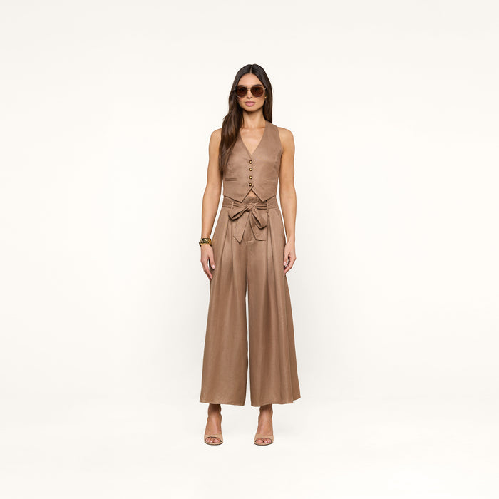 A woman wearing sunglasses and the sleeveless, tan Jianna Vest linen jumpsuit with a waist tie stands against a plain white background, her hands relaxed at her sides, accessorized with heeled sandals and bracelets.