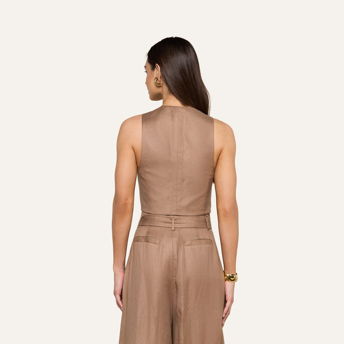 A woman stands with her back to the camera, wearing the sleeveless, light brown Jianna Vest jumpsuit paired with gold hoop earrings and a bracelet, set against a plain light background.