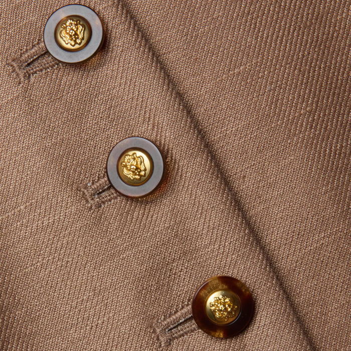 Close-up of the Jianna Vest—a brown cropped vest featuring three round buttons. Each button has a gold center with a raised emblem, encircled by a dark brown ring, and they are sewn in a diagonal line.