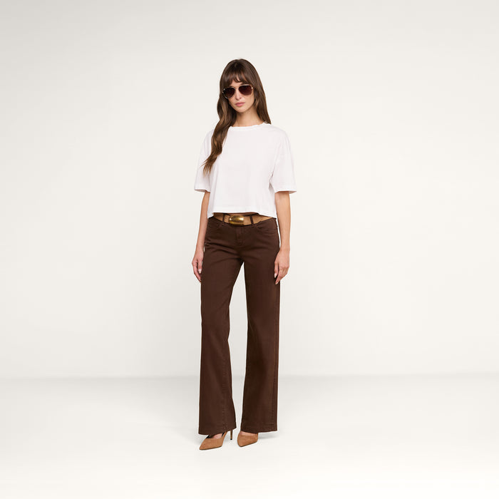 A woman wearing sunglasses, a white short-sleeve top, the Joanne Low-Rise Wide-Leg Jean with a belt, and brown heels stands against a plain white background.