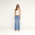 A woman wearing sunglasses, a white tank top, the Joanne Low-Rise Wide-Leg Jean in pinstriped indigo, a brown belt, and sandals stands against a plain white background.
