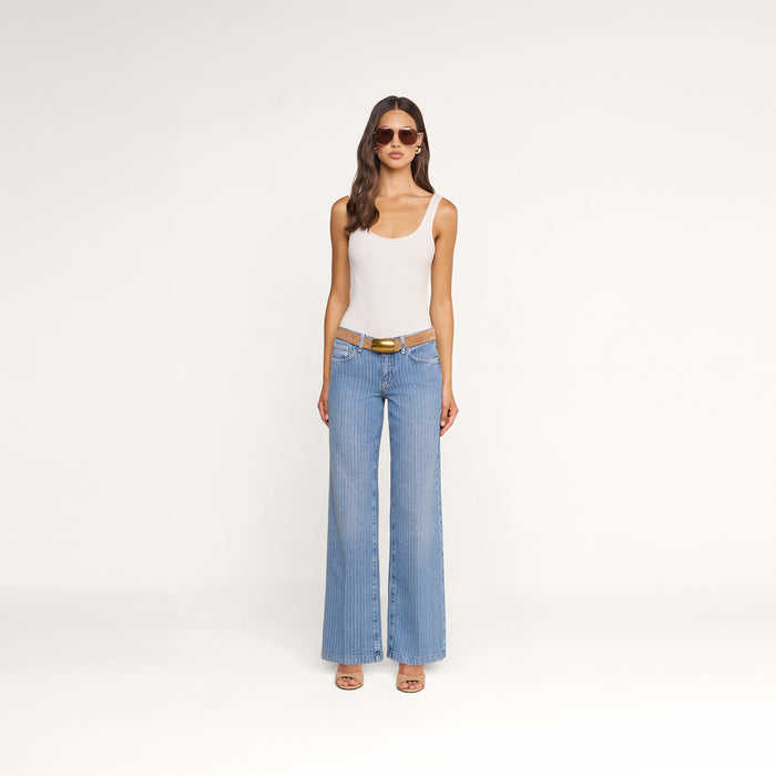 A woman wearing sunglasses, a white tank top, the Joanne Low-Rise Wide-Leg Jean in pinstriped indigo, a brown belt, and sandals stands against a plain white background.