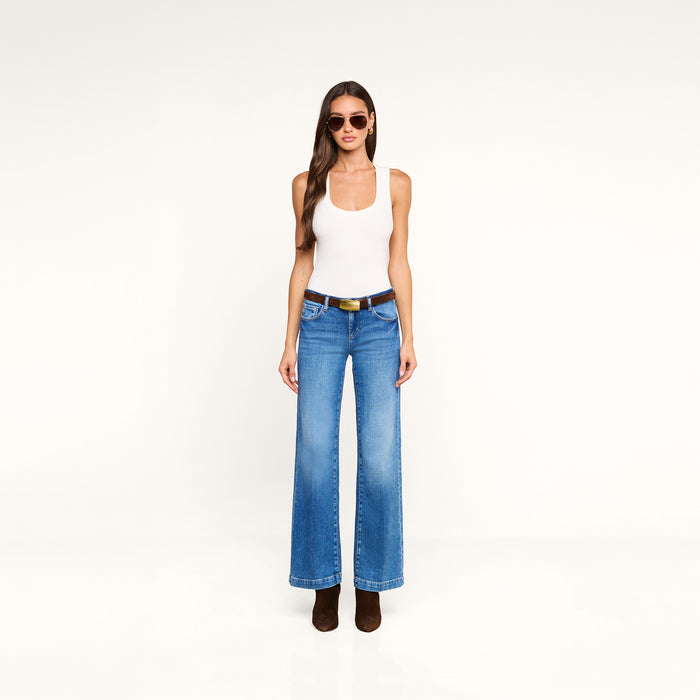 A woman stands against a plain white background, wearing sunglasses, a white tank top, brown boots, and the Joanne Low-Rise Wide-Leg Jean with a black belt. Her outfit features the trendy wide-leg jean styling of the Joanne for a modern look.