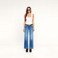 A woman stands against a plain white background, wearing sunglasses, a white tank top, brown boots, and the Joanne Low-Rise Wide-Leg Jean with a black belt. Her outfit features the trendy wide-leg jean styling of the Joanne for a modern look.