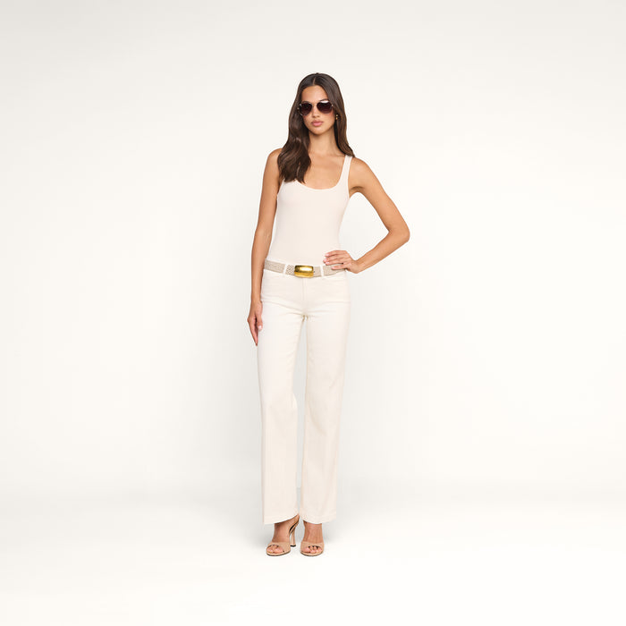 A woman wearing sunglasses, a sleeveless white top, and the Joanne Low-Rise Wide-Leg Jean in an ecru wash with a belt and nude heels stands against a plain white background with one hand on her hip.