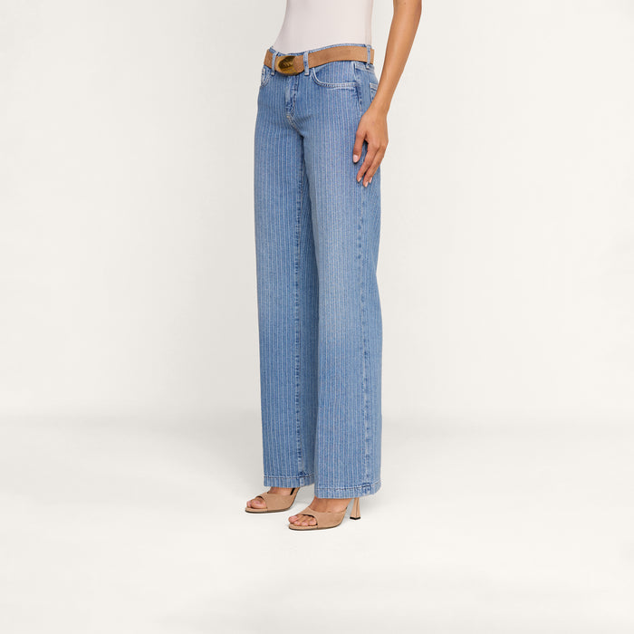 Wearing the Joanne Low-Rise Wide-Leg Jean in pinstriped indigo, styled with a tan belt, a white top, and beige heeled sandals, a person stands against a plain white background.