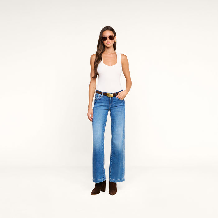 A woman wearing sunglasses, a white sleeveless top, the Joanne Low-Rise Wide-Leg Jean made from stretch denim, a black belt, and brown boots stands against a plain white background.