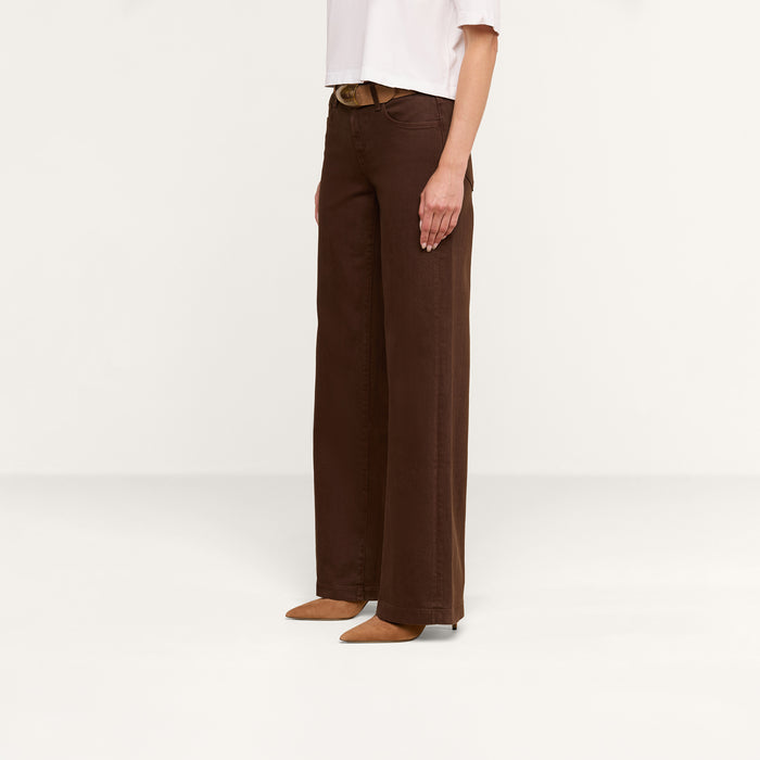 A person stands against a plain white background, wearing a white shirt paired with Joanne Low-Rise Wide-Leg Jeans in dark brown and brown pointed-toe shoes.