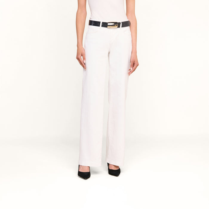 A woman stands against a plain white background wearing the Joanne Low-Rise Wide-Leg Jean in white, paired with a black belt and black heels. Her upper torso is not visible.