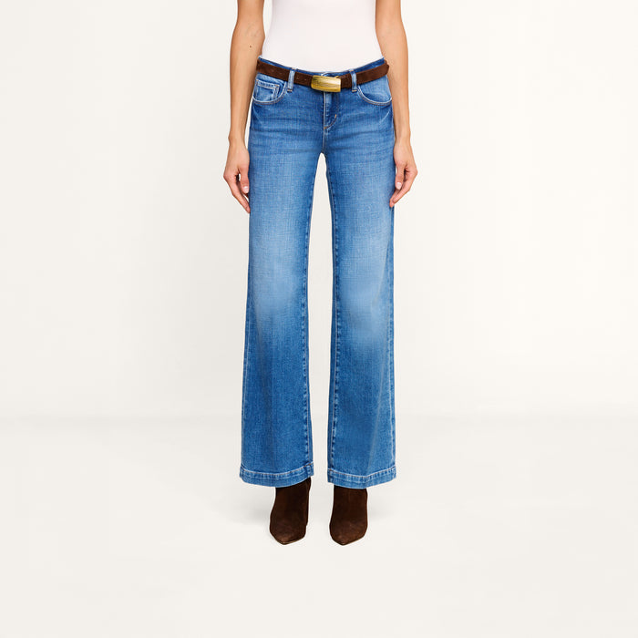 A person wearing the Joanne Low-Rise Wide-Leg Jean in blue stretch denim, styled with a black belt featuring a gold buckle, a white sleeveless top, and brown pointed-toe boots, stands against a plain white background.