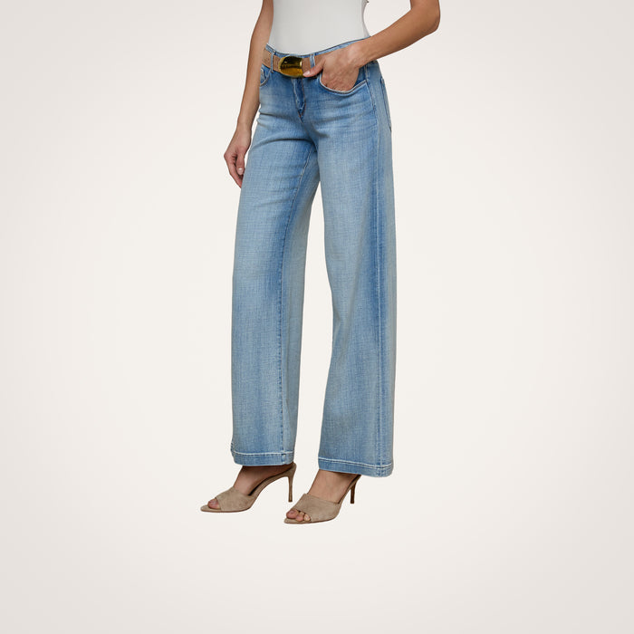 Wearing the Joanne Low-Rise Wide-Leg Jean in light blue stretch denim, a woman pairs them with a white sleeveless top, a brown belt, and beige heeled sandals. With her hands tucked in her pockets, she stands against a plain off-white background.