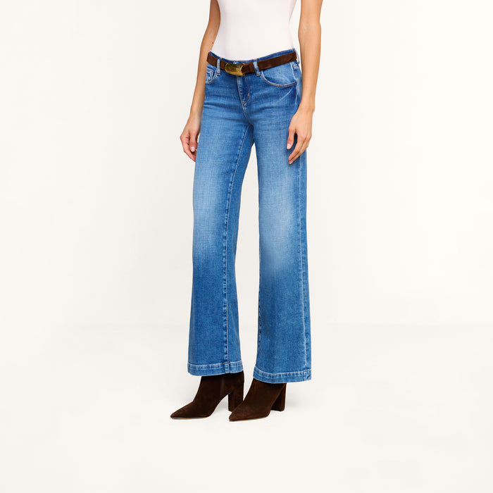 A person wearing a white top and the Joanne Low-Rise Wide-Leg Jean in blue stretch denim, styled with a brown belt and brown heeled boots, stands against a plain white background.