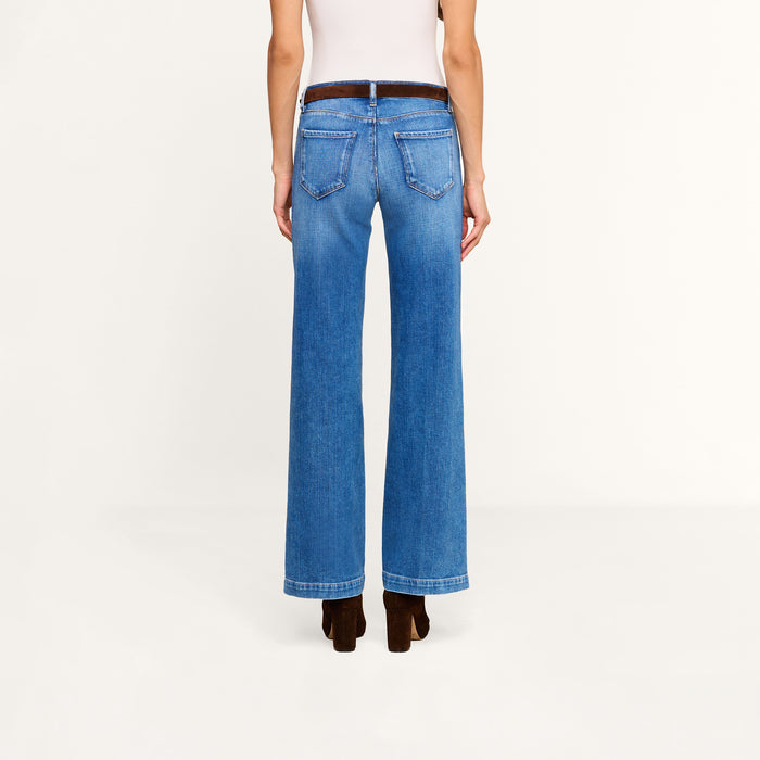 A person stands facing away, wearing the Joanne Low-Rise Wide-Leg Jean in blue stretch denim, a white sleeveless top, and dark brown heeled shoes against a plain, light-colored background.