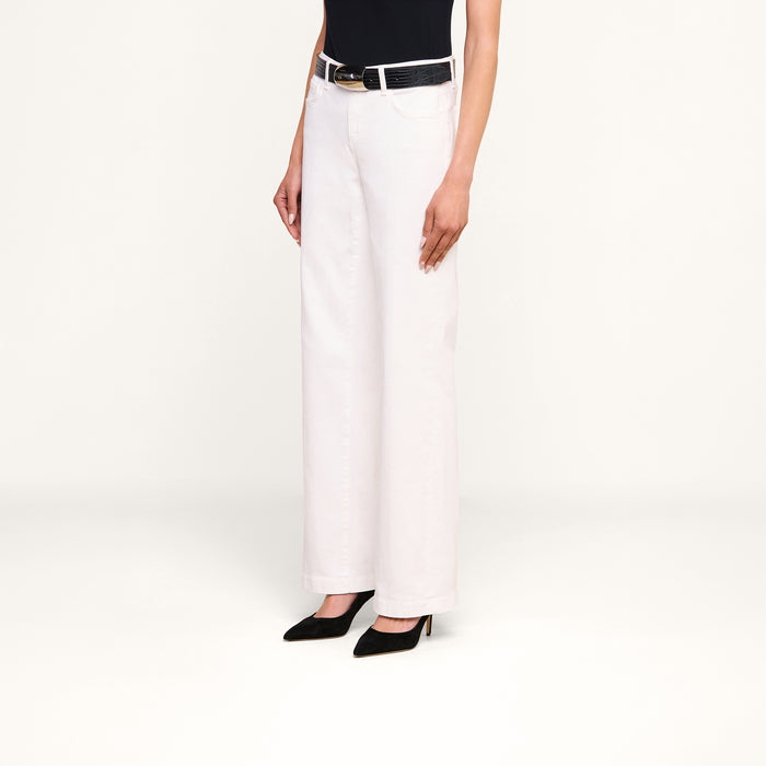 A person wearing the Joanne Low-Rise Wide-Leg Jean in white, paired with a black belt, black sleeveless top, and black heels, stands against a plain white background. The person’s head is not visible in the image.