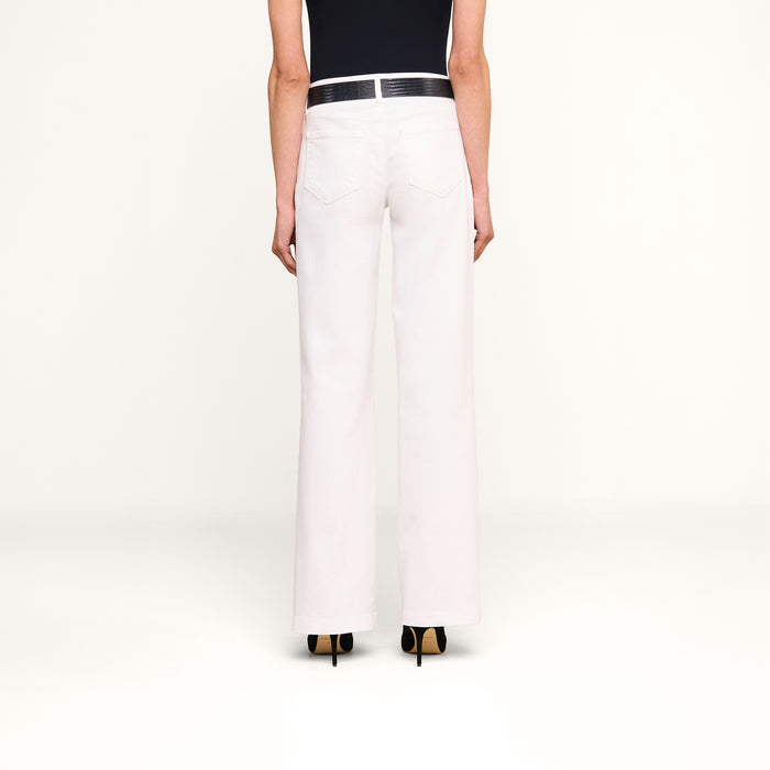 A person stands with their back to the camera against a plain white background, wearing the Joanne Low-Rise Wide-Leg Jean in white, paired with a black belt, a black top, and black high-heeled shoes.