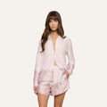 A woman wearing the Judy Blouse, a light pink satin button-down long-sleeve top with a subtle floral pattern, paired with matching shorts, stands against a plain white background.
