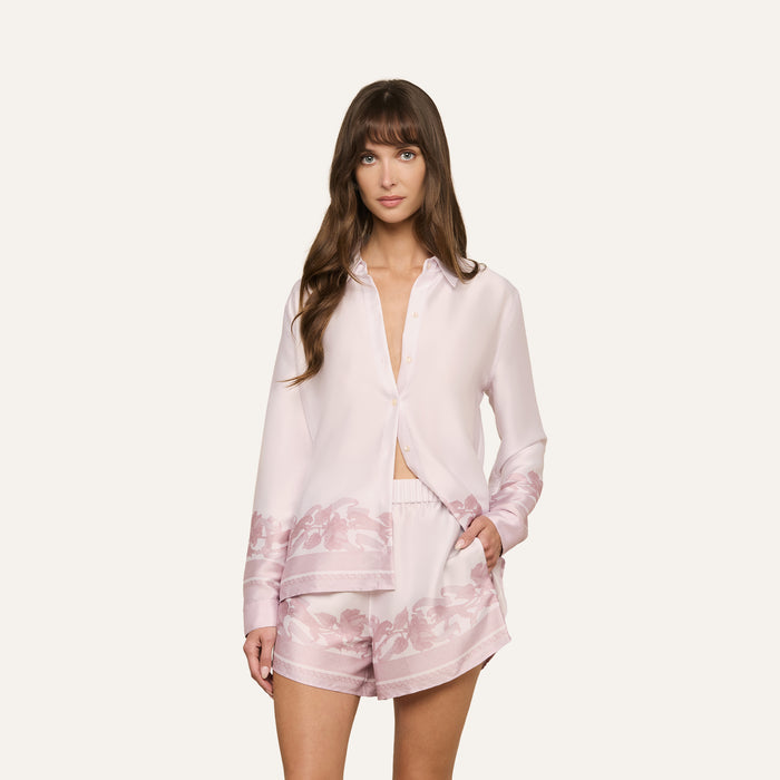 A woman wearing the Judy Blouse, a light pink satin button-down long-sleeve top with a subtle floral pattern, paired with matching shorts, stands against a plain white background.