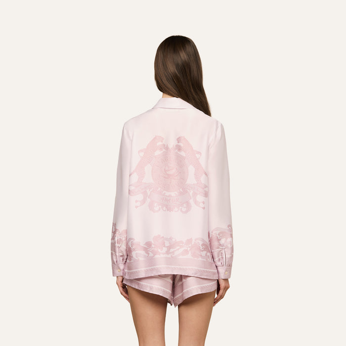 Wearing the Judy Blouse, a woman with long brown hair stands with her back to the camera against a plain, light background. The light pink button-down pajama set features decorative patterns and a crest on the back.