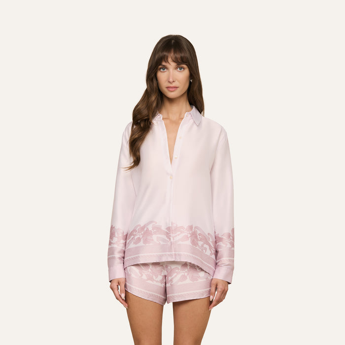 A woman with long brown hair wears the Judy Blouse—a light pink satin button-down shirt with floral patterns near the hem—paired with matching shorts, standing against a plain light background.