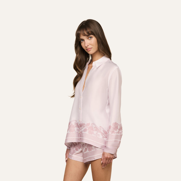 A woman with long brown hair wears the Judy Blouse, a light pink button-down top featuring floral patterns on the hem, paired with matching pink shorts, standing against an off-white background.