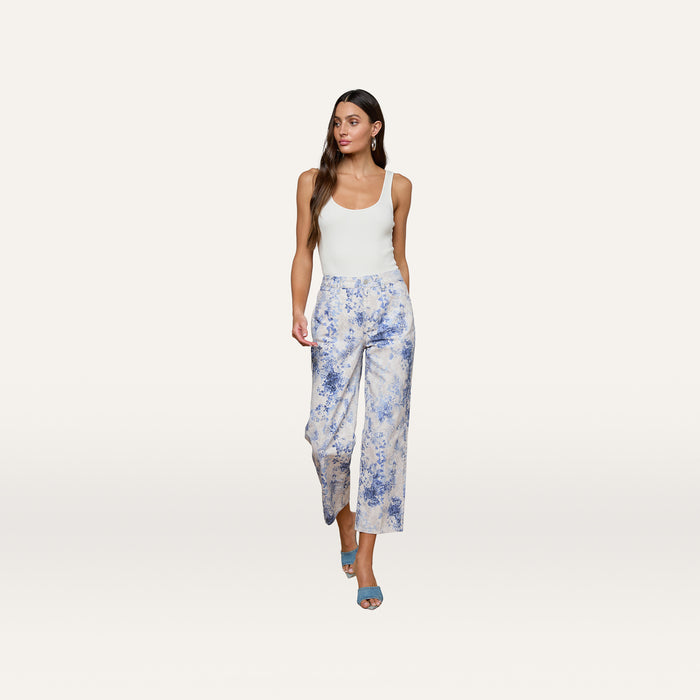 A woman wearing a sleeveless white top and the June Cropped Stovepipe Jean — a high-rise, straight-leg white pant with blue floral paisley print — paired with light blue heeled sandals, stands against a plain background.
