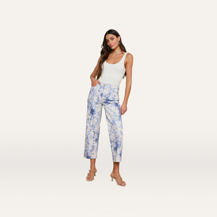 Woman with long brown hair wearing a white tank top, June Cropped Stovepipe Jean in a blue and white floral paisley print, and beige heeled sandals, standing against a plain white background.