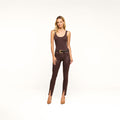A woman stands against a plain white background wearing a sleeveless brown top, a black belt, brown heels, and the Jyothi Coated Jean—a pair of high-rise skinny coated denim pants with ankle slits.