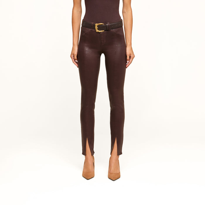 Wearing the Jyothi Coated Jean—high-waisted, brown, and featuring front ankle slits—a person is styled with a black belt with a gold buckle, a sleeveless dark top, and tan pointed-toe heels. The image shows them from the shoulders down against a white background.