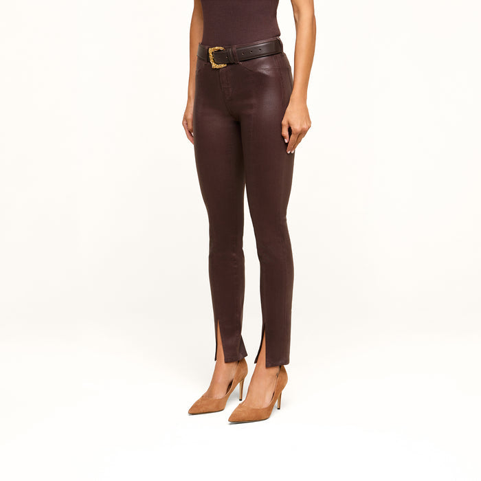 A woman wearing the Jyothi Coated Jean—brown high-rise skinny jeans with front ankle slits—paired with a black top, a black belt featuring a gold buckle, and tan pointed-toe heels, standing against a plain white background.