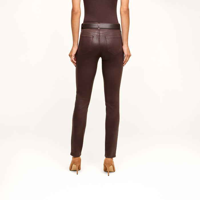 A person stands facing away against a plain white background, wearing a sleeveless brown top, the Jyothi Coated Jean in dark brown—featuring high-rise skinny fit with pockets and an ankle slit—a black belt, and tan high-heeled shoes.