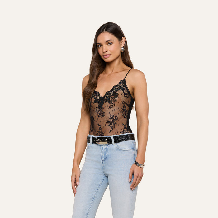 A woman wearing the Kaelith Lace Bodysuit—a black Chantilly floral lace sleeveless design with adjustable straps—paired with light blue jeans, accessorized with earrings, bracelets, and a black belt, stands against a plain white background.