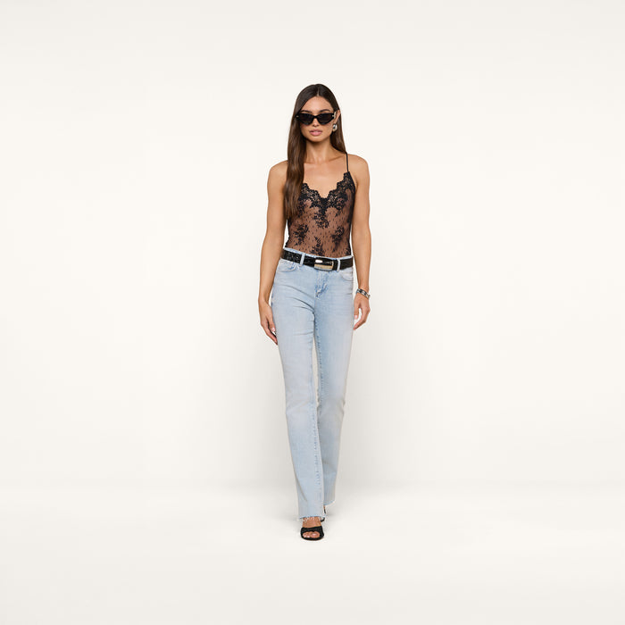A woman wearing sunglasses, the Kaelith Lace Bodysuit featuring sheer Chantilly floral lace and adjustable straps, paired with light blue jeans, a black belt, and black heels, walks confidently against a plain white background.