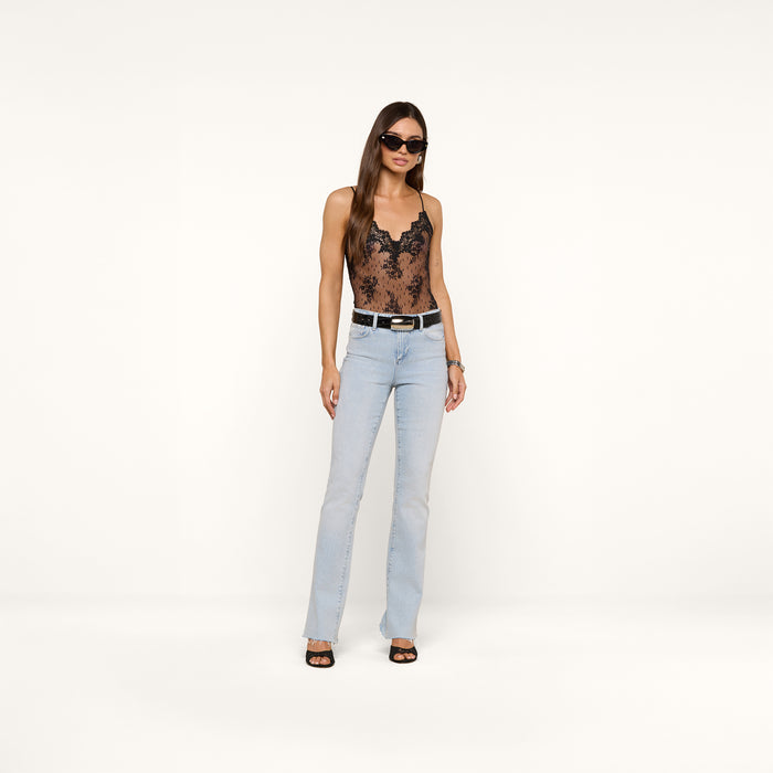 A woman wearing sunglasses, the black Kaelith Lace Bodysuit with adjustable straps, light blue jeans with a belt, and black heels stands against a plain white background.