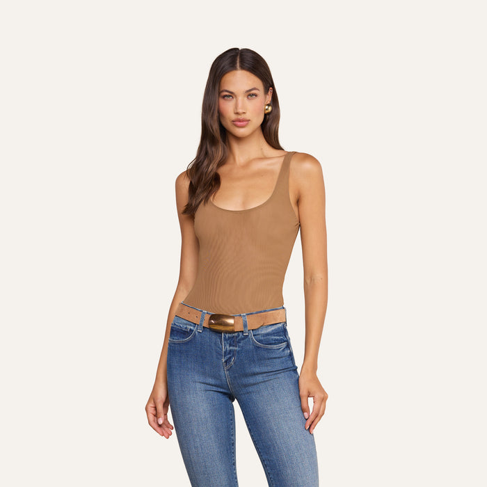 A woman stands facing forward against a plain, light background, wearing the Kalea Mesh Bodysuit in tan as a sleeveless top, paired with blue jeans and a brown belt.