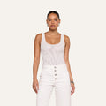 A woman stands facing forward against a plain light background, wearing the Kalea Mesh Bodysuit—a sleeveless white mesh piece—paired with high-waisted white pants featuring silver buttons.