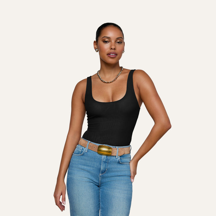 A woman with dark hair pulled back is wearing the Kalea Mesh Bodysuit, styled with a gold necklace, tan belt, and blue jeans. She stands against a plain light background with one hand on her hip.