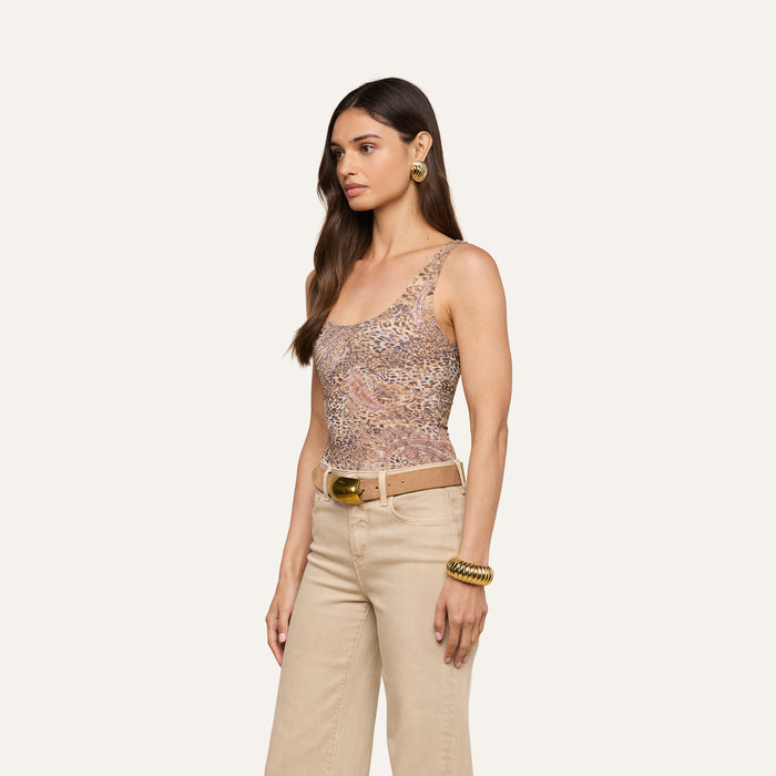Woman standing sideways, wearing the Kalea Mesh Bodysuit—a sleeveless leopard paisley print top—paired with beige belted pants. She has long brown hair, gold earrings, and a chunky gold bracelet. The background is plain and light-colored.