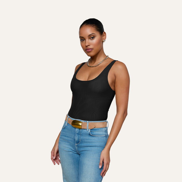A woman stands against a plain background, wearing the Kalea Mesh Bodysuit, blue jeans with a tan belt, and a gold chain necklace. Her hair is pulled back as she looks confidently at the camera.