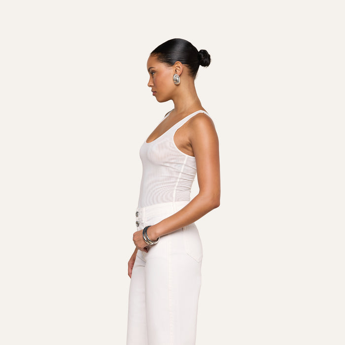A woman stands in profile against a plain light background, her hair in a bun, silver earrings and bracelets accenting her look. She wears the Kalea Mesh Bodysuit in white, styled with matching white pants.