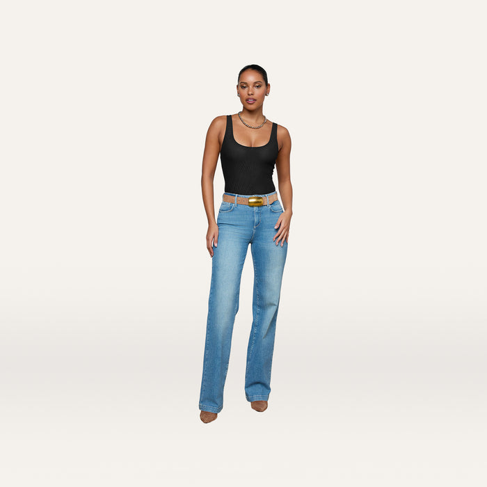 A woman wearing the Kalea Mesh Bodysuit, paired with blue high-waisted jeans and brown heels, stands against a plain white background.